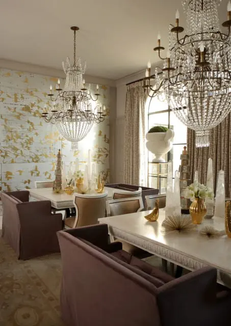 Dining room with two crystal chandeliers, a table with gold decorative objects, and a wall with a gold bird pattern wallpaper.