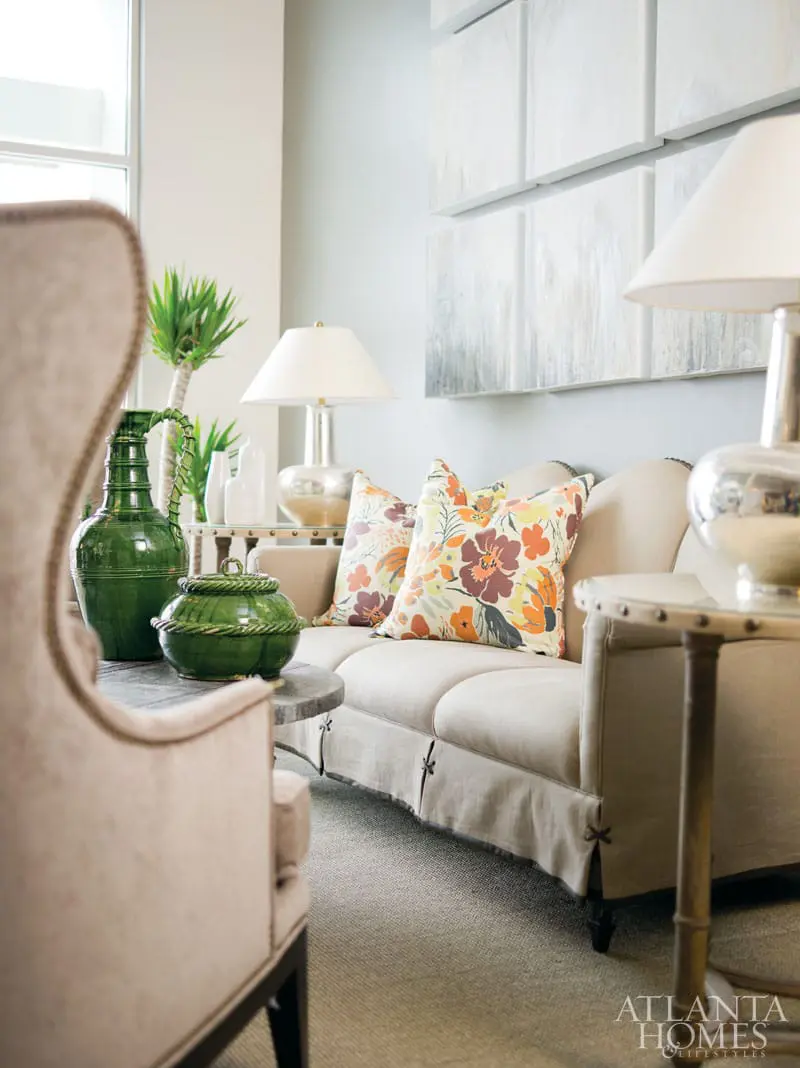Living room with a light beige sofa, floral accent pillows, and a tall green ceramic vase on a side table. Abstract art hangs on the wall.