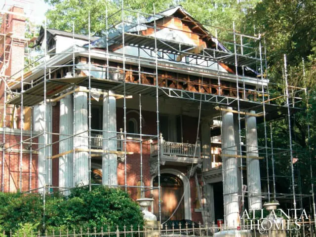 Large brick house with white columns and extensive scaffolding during renovation, surrounded by trees.