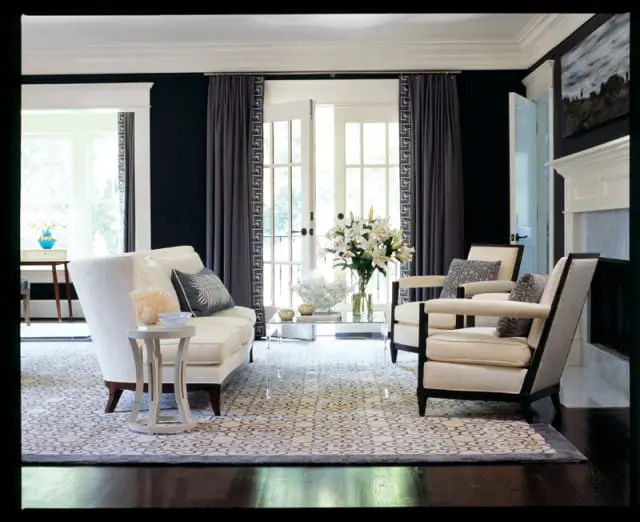 Living room with black walls, white sofa, two cream armchairs with dark wood trim, and a patterned rug. French doors open to a bright outdoor space.
