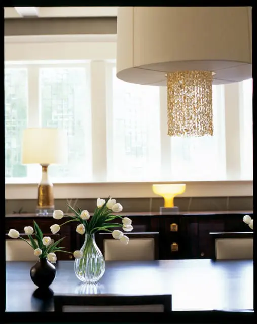 Dining room with dark wood table, upholstered chairs, and a long buffet. White tulips in glass vases sit on the table, beneath a large drum pendant light with a beaded crystal accent.