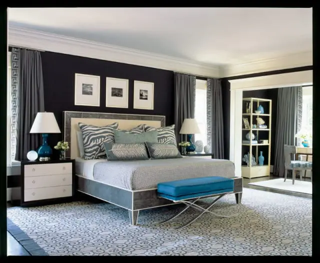 Bedroom with dark walls, a grey and white patterned bedspread, zebra print pillows, and a blue bench at the foot of the bed. A white dresser with a blue lamp sits beside the bed.