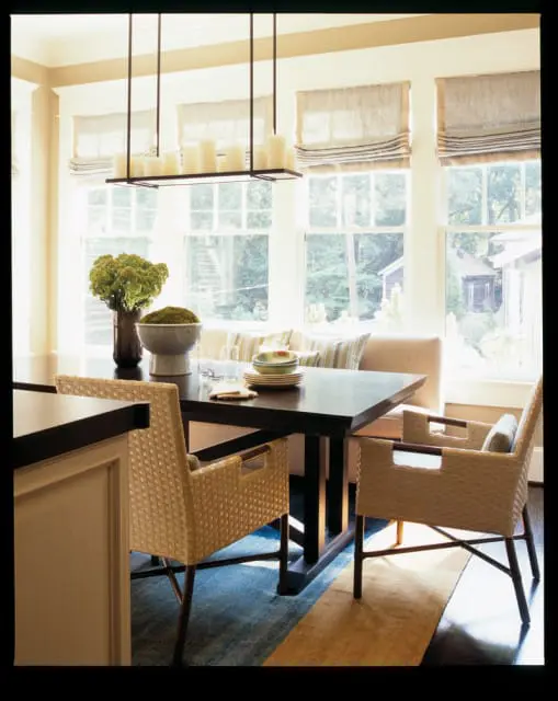 Dining area with dark wood table, woven chairs, banquette seating, and a modern candelabra chandelier. Large windows overlook a wooded backyard.