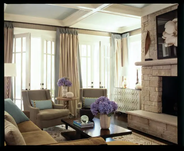 Living room with coffered ceiling, stone fireplace, upholstered armchairs, and a dark wood coffee table with floral arrangements.
