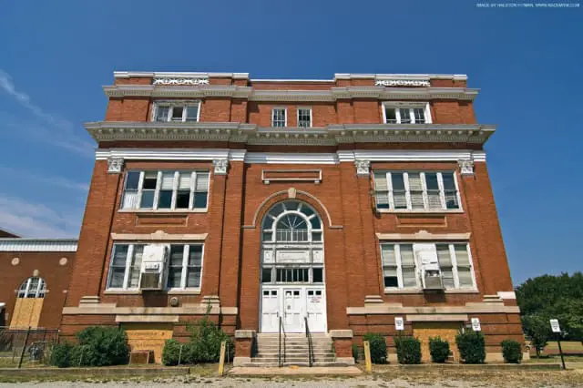 Red brick building with white trim, arched window above double doors, and boarded-up lower windows under a clear blue sky.
