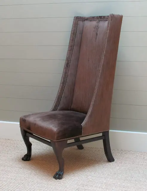 Tall wingback chair upholstered in brown textured fabric with nailhead trim, dark wood frame, and carved paw feet, set on a neutral woven rug against a pale green paneled wall.