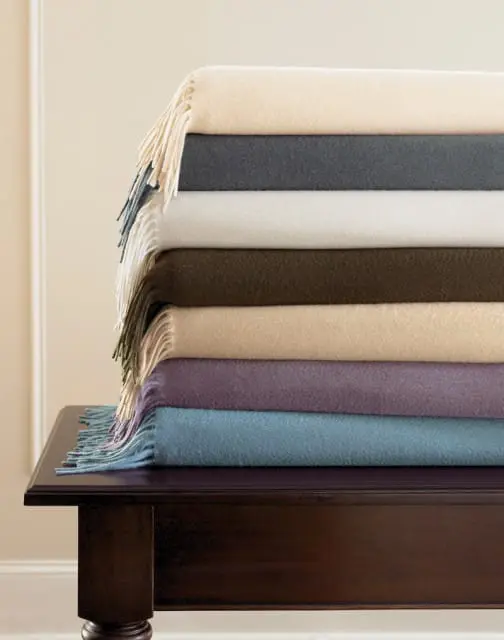 Stack of seven folded throws in cream, charcoal, white, olive, tan, plum, and blue, with fringe details, resting on a dark wood surface.