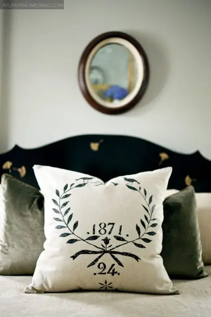 Bedroom with a black scalloped headboard, a cream pillow with a black laurel wreath and '1871. 24.' stencil, and two dark green velvet pillows.