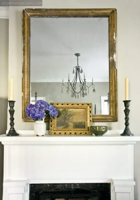 Mantelpiece with a large antique gold-framed mirror reflecting a crystal chandelier, flanked by candlesticks and a vase of purple hydrangeas next to a landscape painting and a green bowl.