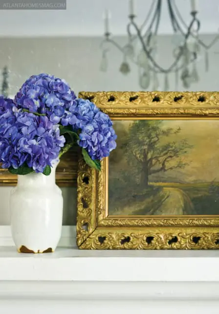 White ceramic vase with blue hydrangeas next to a landscape painting in an ornate gold frame on a white mantelpiece.