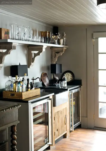 Wet bar with a stainless steel sink, granite countertops, and two wine refrigerators. A wicker tray holds various liquor bottles. Shelves above display glassware and barware.
