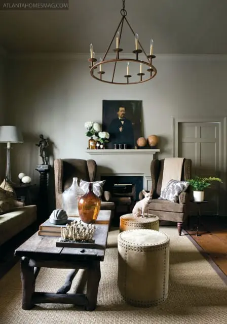 Living room with taupe walls, a dark wood coffee table, two brown wingback chairs, a fireplace with a portrait above it, and a small dog sitting on an ottoman.