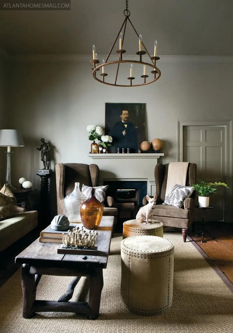Living room with taupe walls, a dark wood coffee table, two brown wingback chairs, a fireplace with a portrait above it, and a small dog sitting on an ottoman.