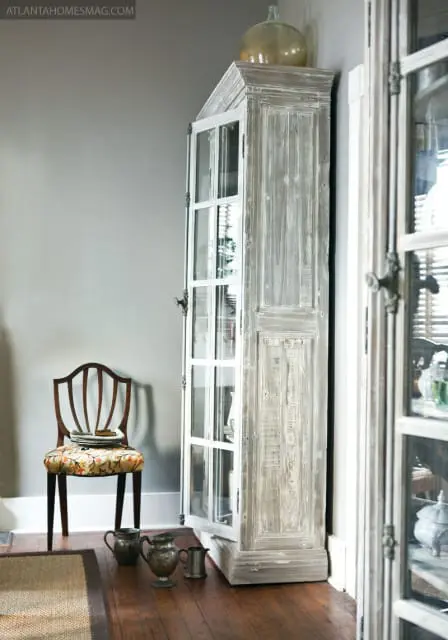 Corner cabinet with glass doors and distressed wood finish, next to a wooden chair with floral upholstery. Antique metal pitchers sit on the wooden floor.
