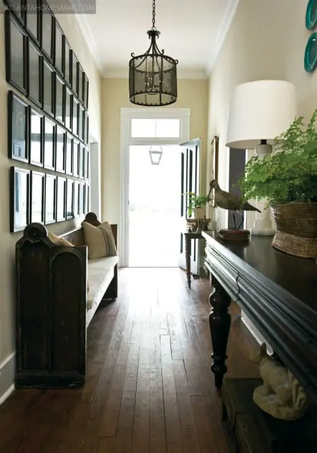 Hallway with a wall of framed art, a dark wood bench, a large dark wood console table with a lamp and plant, and a chandelier above a doorway.