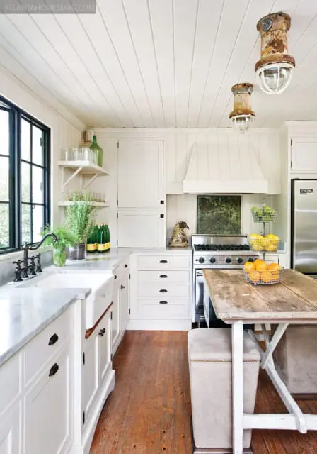 White kitchen with white paneled ceiling, farmhouse sink, marble countertops, white cabinets, stainless steel range, and a rustic wooden island with upholstered stools.