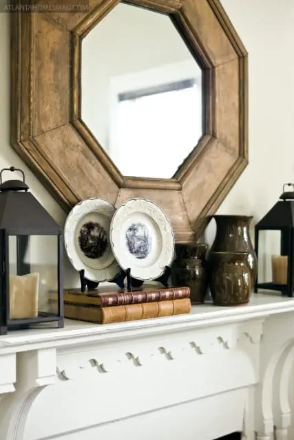 White mantelpiece with an octagonal wooden mirror, two decorative plates, stacked antique books, brown ceramic vases, and black lanterns with candles.