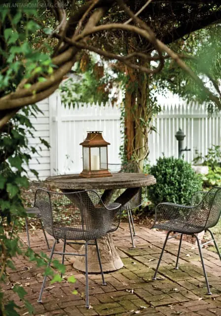 Stone patio table with metal chairs and a lantern, surrounded by greenery and a white picket fence.