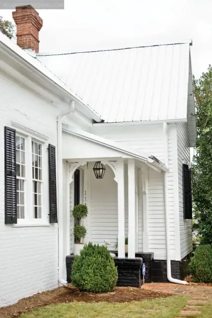 White brick house with black shutters, a white metal roof, and a covered porch with a black lantern.