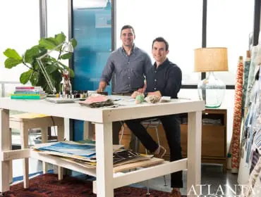 Two men stand behind a white work table in a studio, with art supplies, books, and plants. Rolls of fabric are visible on the right.