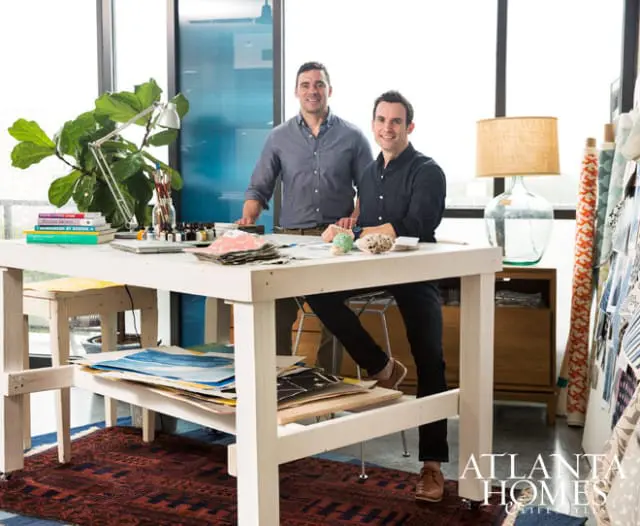 Two men stand behind a white work table in a studio, with art supplies, books, and plants. Rolls of fabric are visible on the right.