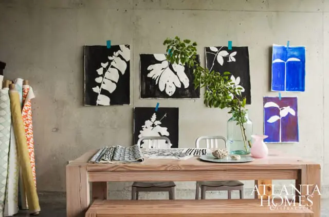 Wooden table with chairs in front of a concrete wall displaying various art pieces and rolls of fabric.