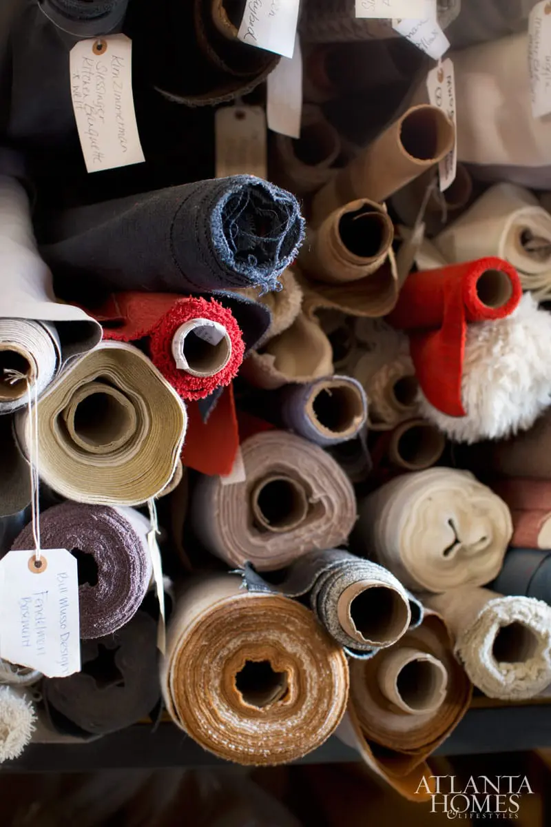 Close-up of fabric rolls in various colors and textures, including navy, red, beige, and brown, with some rolls tagged with handwritten labels.