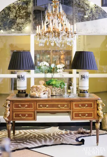 Antique wooden desk with gold accents and ornate drawer pulls, topped with two black pleated lamps, orchids, and decorative sculptures, beneath a crystal chandelier and distressed mirror wall.
