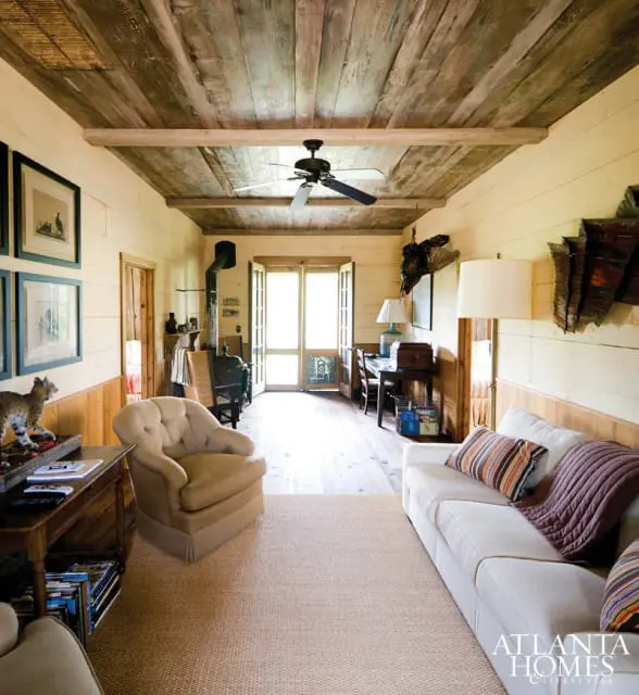 Living room with rustic wood-paneled walls and ceiling, a white sofa with striped pillows, a tan armchair, a wood-burning stove, and a desk area.