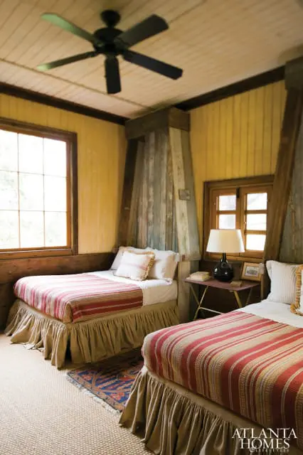 Bedroom with two twin beds featuring red and white striped comforters, tan bed skirts, and a black ceiling fan. Yellow paneled walls and rustic wooden beams add to the room's charm.