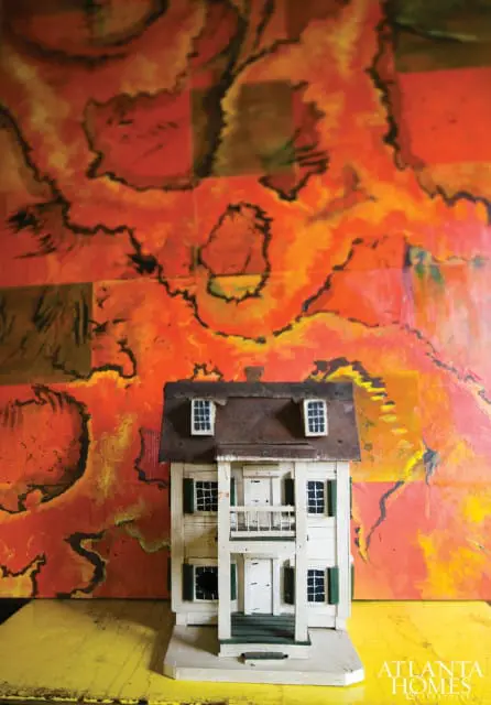 Dollhouse replica of a white colonial-style house with green shutters and a balcony, set against a vibrant orange and black abstract painting.