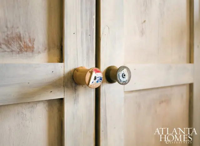 Close-up of two wooden cabinet doors with a distressed white finish and unique drawer pulls. One pull is a wooden spool with a blue and red graphic, the other is a metal knob with a circular graphic.