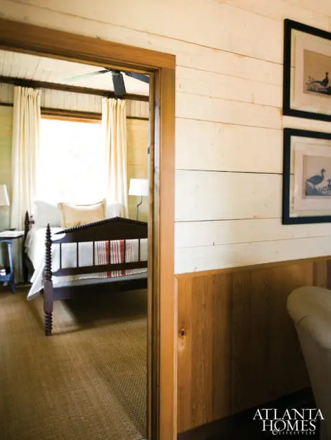 Bedroom with a dark wood spindle bed, white linens, and cream curtains, viewed through a doorway. The room has a ceiling fan and is carpeted with a natural fiber rug.