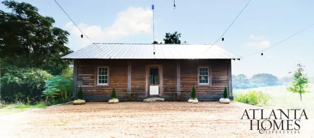 Wooden cabin with a metal roof, two windows, and a central door, set in a grassy field with trees and a logo for Atlanta Homes & Lifestyles in the lower right corner.