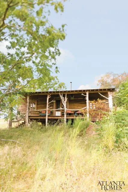Rustic wooden cabin with a covered porch, surrounded by tall grass and trees under a bright blue sky.