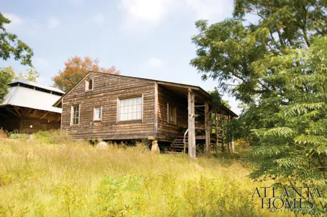 Rustic wooden cabin with a covered porch, set on a grassy hill with trees and a barn in the background.