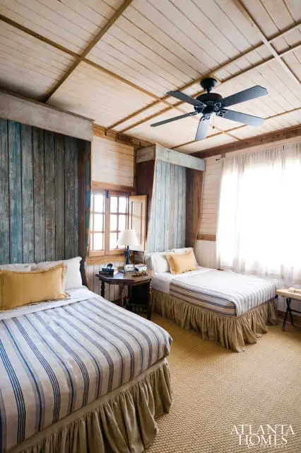 Bedroom with two twin beds featuring blue and white striped comforters, tan bed skirts, and yellow accent pillows. Walls are paneled wood with accents of distressed blue wood. A black ceiling fan is visible.