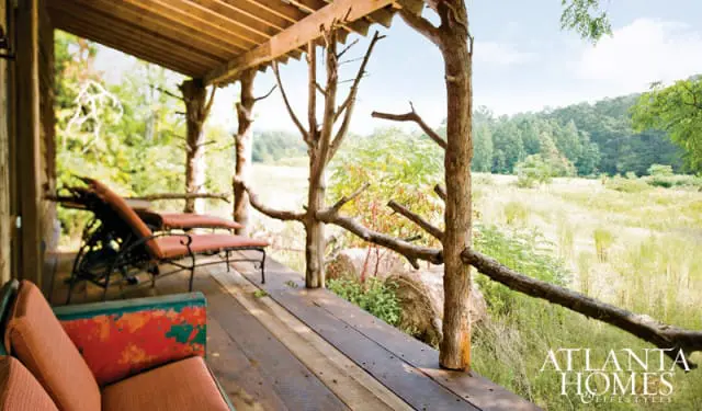 Rustic wooden porch with natural tree branch railings and outdoor seating overlooking a grassy field and forest