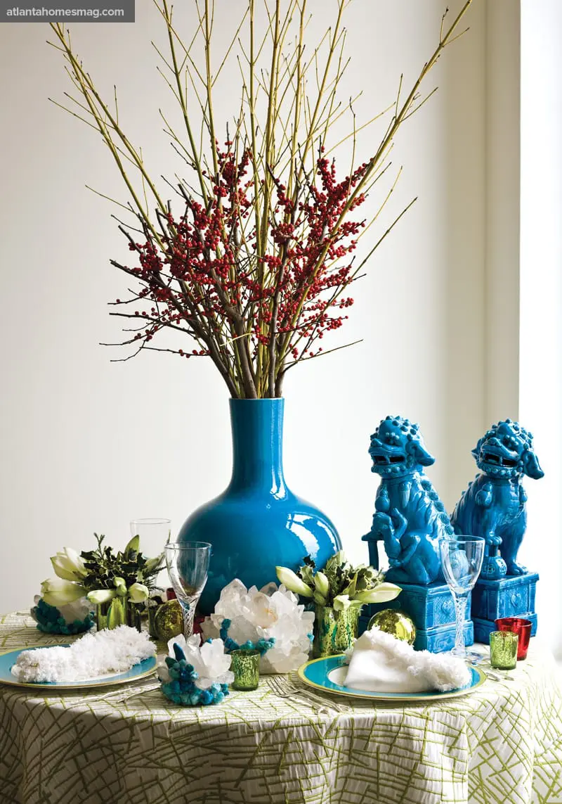 Table set with a green and white patterned tablecloth, blue vase with red berries and branches, two blue foo dogs, crystal clusters, and place settings.