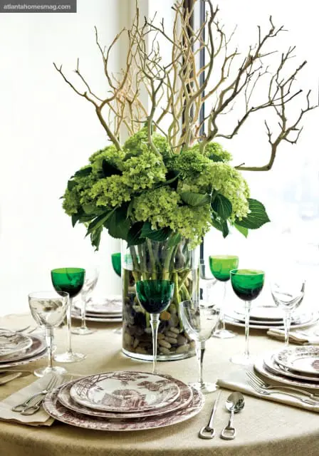 Dining table set with toile pattern plates, clear and emerald green wine glasses, and a tall vase filled with green hydrangeas and branches.
