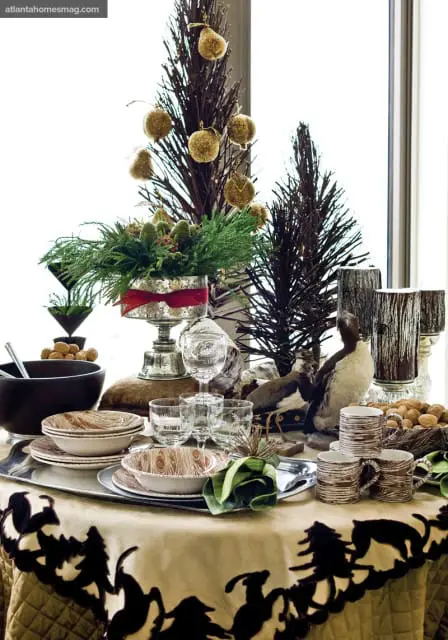 Table setting with stacked plates and bowls featuring wood grain pattern, crystal glassware, stuffed birds, and decorative twig trees with gold ornaments.