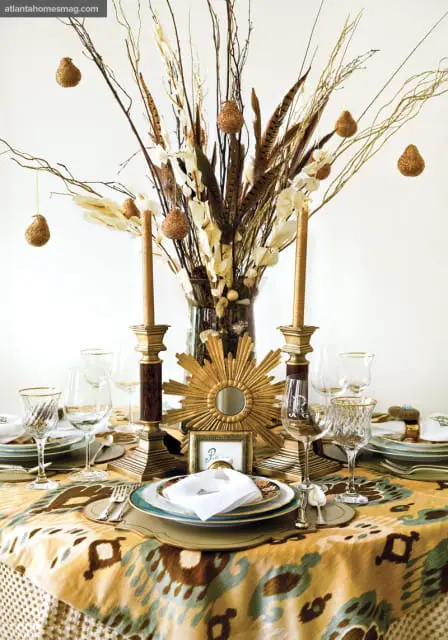 Dining table set with a patterned tablecloth, gold candlesticks, crystal glassware, and a centerpiece of dried branches, feathers, and glittered ornaments.