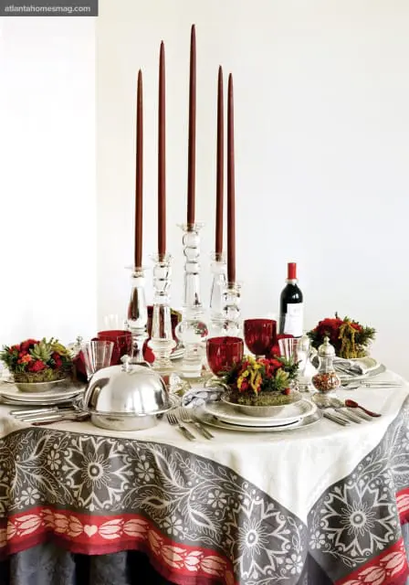 Round dining table set with white tablecloth featuring a gray and red floral border, crystal candlesticks with tall red candles, red glassware, floral centerpieces, and a silver cloche.