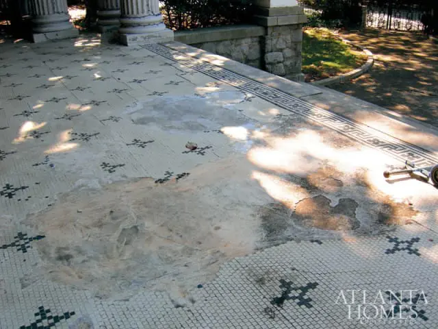 Close-up of a mosaic tile floor with a Greek key border and scattered dark floral patterns, showing areas of wear and damage.