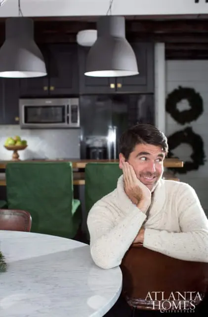 Man in cream cable-knit sweater with shawl collar resting his chin on his hand, looking to the right. Dark kitchen cabinets and green chairs visible in background.