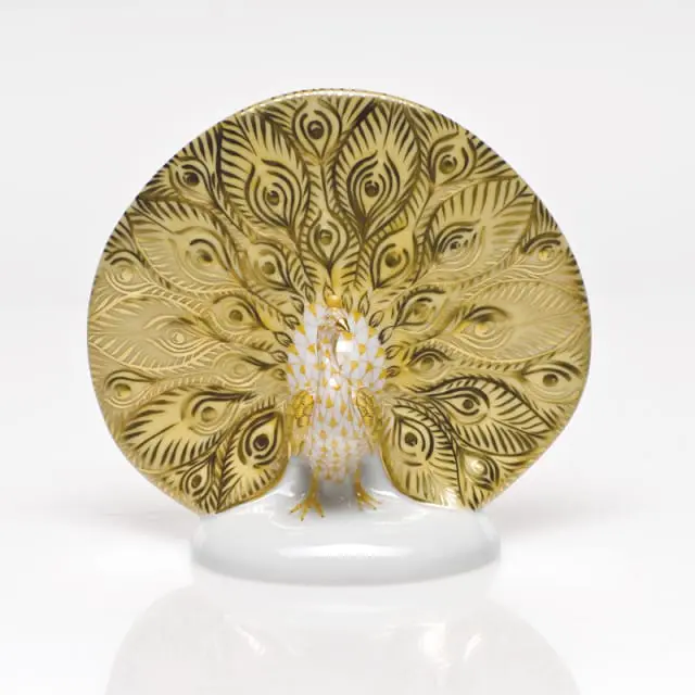 Porcelain peacock figurine with fanned tail feathers decorated in gold and white patterns, standing on a white base.