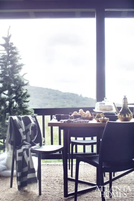 Dining table set with coffee cups, pastries, and a cake under a glass dome, with chairs draped in a geometric blanket, overlooking a mountain view.