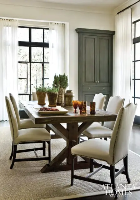Dining room with a rustic wooden table, upholstered chairs, and a tall gray cabinet. Large windows with sheer curtains are visible.