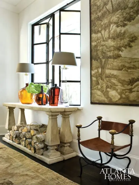 Interior scene with a console table displaying amber glass vases and green floral arrangement, a stack of firewood below, and a metal chair with leather seat next to a large window.