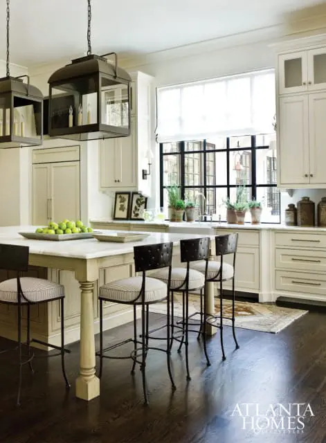 Cream kitchen with marble island, dark wood floors, black and white striped bar stools, and large black-paned window with potted plants.